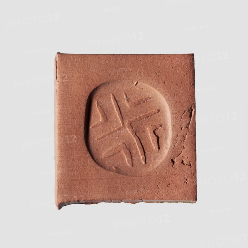Stamp seal with geometric ornament. A prism-shaped stamp seal with rounded edges and an oval, nearly rectangular base. A hole is drilled through the longer diagonal side walls transverse to the longitudinal axis of the stamp. On a flat base is carved a shallow geometric ornament consisting of regularly spaced, slightly open hooks: two large hooks with their tips touching in the center and four small hooks in triangular fields between the arms of the large hooks. Materials: seal. Culture/Date: Halaf culture, c. 6th–5th millennium BC ca. 6000–5000 BC. Place of origin: Northern Mesopotamia Iraq/Syria/Turkey. Dimensions: height 0.8 cm; width 1.5 cm; depth 1.7 cm; hole diameter 0.2 cm. Creator: unknown., A square piece of reddish-brown clay or terracotta with a circular impression in the center containing a simple carved cross-like pattern, sitting on a light gray background; the square shows rough edges and small surface cracks and texture, with visible variations of reddish-brown and subt...