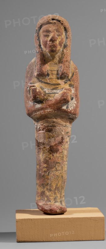 Ushabti, Restitution. Hieroglyphs; funerary figurine; wig; hoes; grave goods; mummy-shaped figurine. Arms crossed on the chest, right folded over left, holding hoes in each hand. Rectangular basket slung over both shoulders on the back. Painted tools. Wide painted usekh necklace on the chest; bracelets on the wrists. Long three-part wig, ears uncovered. Round face with large eyes, wide nose, mouth barely visible; wig and facial details painted. Painted inscription around the figurine's legs, mostly erased; text illegible. Circa 1295–1186 BC; New Kingdom, 19th Dynasty, Egypt. Shabti. Height 15.1 cm; width 4.2 cm., A weathered small statue of a human figure with crossed arms, standing on a rectangular base, showing a rounded head and a stylized wig or hair that frames the face; the surface has worn areas and textured pitting. The visible colors include reddish-brown, muted yellow, pale beige, black accents, and traces of white, with the base in a tan or light brown hue. The figure's tors...