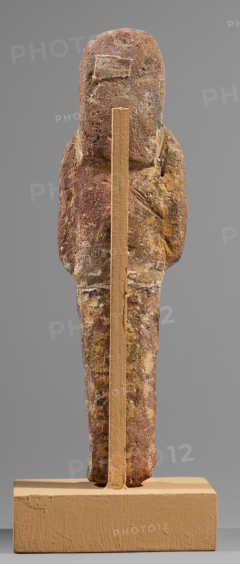 Ushabti, Hieroglyphs, funerary figurine mummy-shaped shabti, c. 1295–c. 1186 BC, New Kingdom, 19th Dynasty, Egypt. Height 15.1 cm, width 4.2 cm. Arms crossed on the chest, right over left, holding hoes in each hand. Rectangular basket slung over both shoulders on the back with painted tools. Wide painted usekh necklace on the chest, bracelets on the wrists. Long three-part wig leaving ears uncovered. Round face with large eyes, wide nose, mouth barely visible; wig and facial details painted. Painted inscription around the legs, mostly erased and illegible., A roughly human-shaped carved object mounted on a vertical support and rectangular base is visible, showing a worn surface with rough texture and irregular patches; the object displays a palette of brown, reddish-brown, yellow, and small areas of whitish tones, while the support and base are a uniform beige color; the shape includes a rounded top, slightly broader upper section suggesting shoulders, a narrower lower section, and vis...
