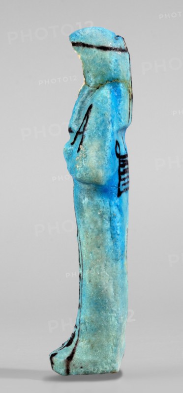 Amenhotep's shabti with baskets on the back; hieroglyphs; headbands; wigs; baskets; hoes; grave goods; mummy-shaped figurine. Arms crossed on the chest, the right folded over the left; hoes in both hands. On the back is a rectangular basket with loops. Painted tools. Three-part, long, smooth wig with a headband; ears covered; face damaged; painted headband. Painted inscription in one column on the front of the legs reading: Osiris the father of God ... scribe of the temple of Amun-Re, Amenhotep. Ca. 1069–715 BC, Third Intermediate Period, 21st–23rd Dynasty, Egypt. Shabti; height 13.3 cm, width 4.2 cm., A small vertical figurine viewed in profile with a rounded head and elongated body, predominantly bright turquoise and blue with areas of lighter greenish-blue patina, showing a few narrow dark lines and markings on the head, torso, and lower leg, a slightly bulbous midsection suggesting a stylized torso, a hint of a raised ear or headdress at the back of the head, a gently curved foot a...