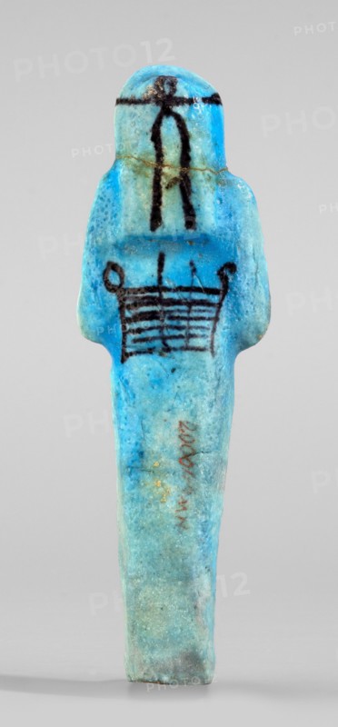Amenhotep's shabti with baskets on the back; arms crossed on the chest, right folded over left, holding hoes in both hands; rectangular basket with loops on the back; painted tools. Three-part long smooth wig with headband covering ears; face damaged; painted headband. Painted inscription in one column on front of legs reading Osiris the father of God ... scribe of the temple of Amun-Re, Amenhotep. Ca. 1069–715 BC, Third Intermediate Period, 21st–23rd Dynasty, Egypt. Shabti, height 13.3 cm, width 4.2 cm., A small figurine-shaped object stands upright against a neutral background, predominantly bright turquoise and light blue with areas of pale green and hints of darker blue; a few black painted linear motifs appear on the upper body and chest, and there are traces of brownish discoloration and tiny flecks of gold on the surface; the form has rounded shoulders, a bulbous head, and an elongated narrow lower body with a slightly irregular, textured surface.