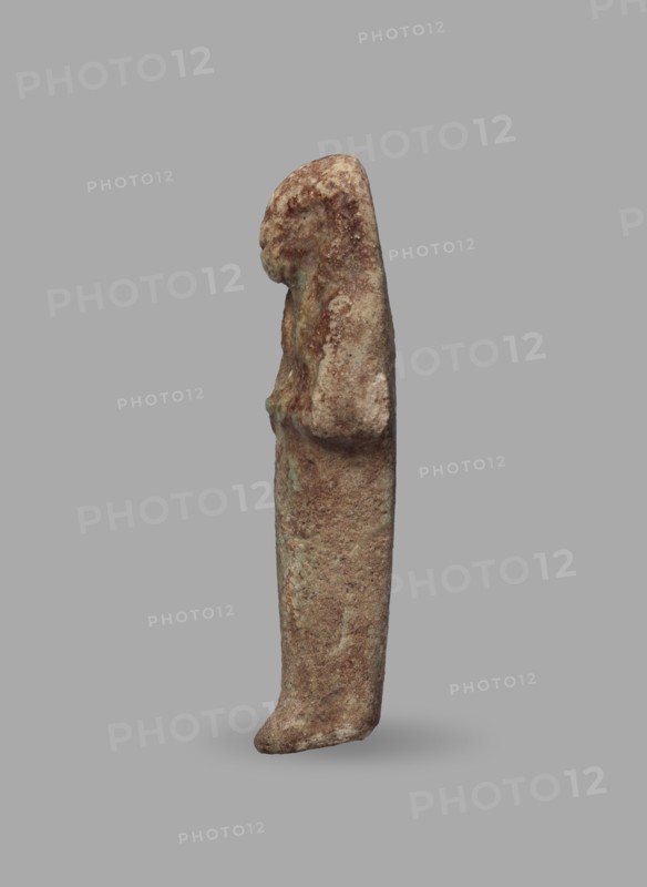 Ushabti, Mummy-shaped funerary figurine, ushabti. Arms crossed on the chest, hands arranged in a line next to each other, holding hoes in both hands with slightly convex tools. Long, three-part smooth wig leaving ears uncovered. Round face with illegible facial details. Not inscribed. ca. 1069–715 BC; Third Intermediate Period, 21st–23rd Dynasty, Egypt. Height 5.5 cm; width 1.8 cm., A small vertical statue of a standing figure shown in profile against a plain gray background, with a textured surface in shades of brown, tan, and hints of green; the figure has a rounded head and a long, narrow body with subtle raised areas suggesting folded arms and a gently protruding foot, the surface appearing weathered and mottled with lighter and darker brown patches and small greenish spots.