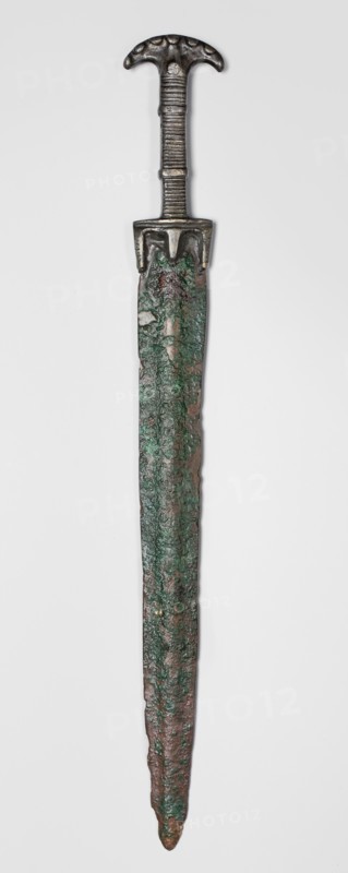 Double-edged sword, Bosporan workshop. Short sword, cast in one piece. Double-edged blade with a centric point and a prominent bulge running through the center of both faces. Hilt divided into three segments: a crescent-shaped pommel, decorated on both sides with small oval knobs imitating rivet heads; one such knob at the transition to the hilt shaft on each side. b square-section shaft decorated on two opposite sides with horizontal incisions; shaft is encircled in two places by a convex, flat band; guard at base of blade. c guard in the form of a horizontal profiled strip from which three protrusions emerge — two longer along the blade line and one shorter on the axis extension. 6th century BC, Black Sea region Bosporan, weapon. Height 45.3 cm; width 5.6 cm; depth 1.7 cm; length 45.5 cm; weight 0.566 kg., An elongated metal object with a dark metallic handle and a rounded, T-shaped pommel showing patterned indentations, a ridged grip, and a short guard; below the guard a long narrow...