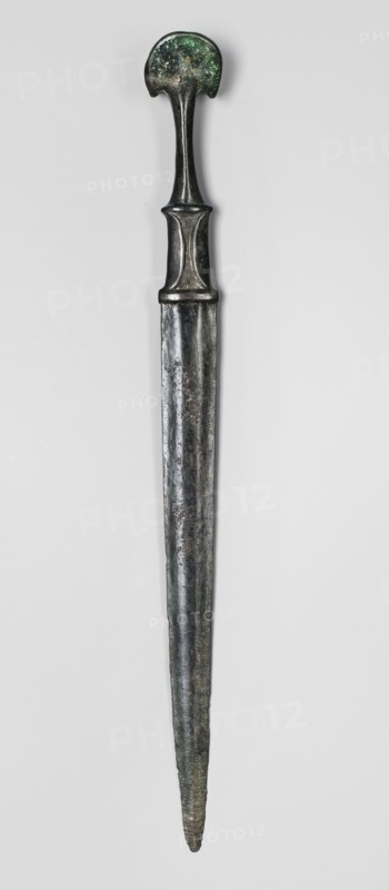 Dagger, unknown Luristan workshop. A fully cast Luristan bronze dagger with a full hilt and a flared pommel. The pommel, shaped like two angled, opposing crescent-shaped lobes, merges into a narrow shaft that continues into a rectangular element imitating a casing with curved mounting plates at the bottom of the hilt such hilts also had casings of various materials applied. The hilt merges directly into a double-edged blade that tapers evenly toward the tip. A ridge runs from the tip of the engraving toward the center of the superstructure, which after a short distance splits, creating a long, narrow, triangular flat field. 9th–6th century BC; Iron Period II. Luristan, Iran. Weapon. Height 35.7 cm; width 3.2 cm; depth 3.4 cm; weight 264 g., A long metallic object with a rounded top that has a mottled green and dark patina, a narrow curved neck leading to a wider midsection with subtle raised ridges, and a tapering blade-like lower portion showing a dark silvery and greenish surface wit...