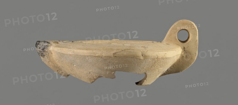 Oil lamp with an image of Helios. Roman single-burner oil lamp made from a mold. Round, flat container with smooth, flat arms; one arm has an ingot. Vertical vice opposite the burner decorated with two grooves. Round, slightly concave discus bounded by three concentric grooves; center shows a bust of Helios. Next to the bust is a round olive hole made after the lamp was pressed from the mold. Burner short, incorporated into the arms, round Bussière type 2b, blackened. Bottom and part of the burner are broken. Light clay may indicate manufacture in Corinth Bernhard suggests an eastern workshop. 1st century., A broken shallow bowl or lamp-shaped ceramic object in beige and tan tones with a rounded rim and a curved hooked handle on one side that has a circular hole, a darker worn tip on the opposite edge showing black and gray discoloration, rough textured surfaces with chips and missing fragments along the lower edge, and a neutral gray background.