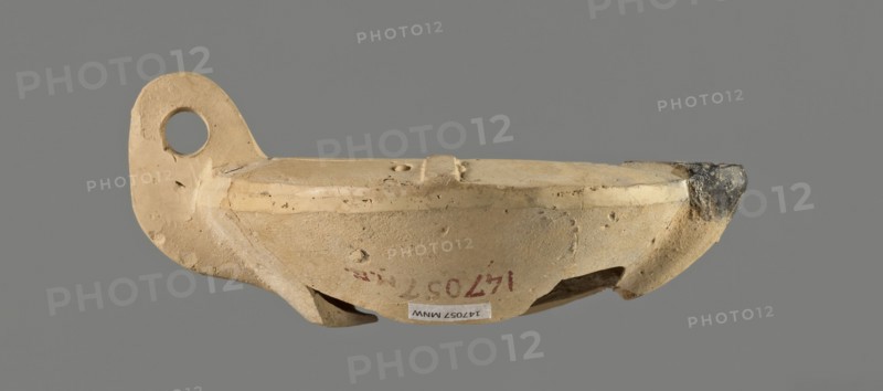 Oil lamp with an image of Helios. Single-burner Roman oil lamp, Broneer Lamp type XXVII C, made from a mold. Round, flat container with smooth, flat arms; one arm has an ingot. Vertical vice opposite the burner decorated with two grooves. Round, slightly concave discus bounded by three concentric grooves; center bears a bust of Helios. Next to the bust is a round olive hole made after molding. Burner short, incorporated into the arms, round Bussière type 2b, blackened. Bottom and part of the burner are broken. Light clay may indicate production in Corinth Bernhard suggested an Eastern workshop. 1st century; oil lamp., A small beige clay vessel with a rounded body and a raised rim, showing rough and pitted surface texture and a few chips and cracks; one end has a flat, ear-like extension with a circular hole, and the opposite rim end is darker with soot-like dark gray discoloration; the vessel rests against a neutral gray background and displays subtle variations of beige, tan, light br...