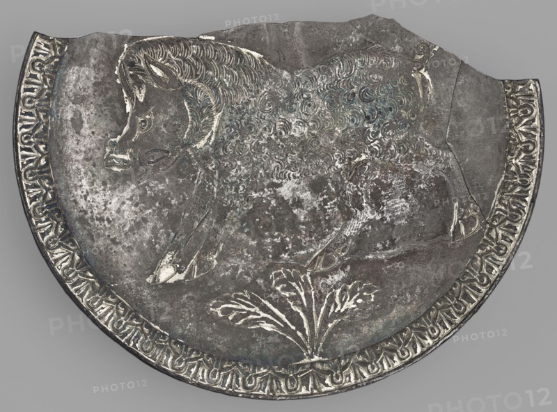 Platter with a depiction of a boar, Byzantine workshop, boar iconography, platter, 2nd half of the 5th century AD, found in Toulouse, Roman Empire 27 BC–476, vessel, weight 505.81 g, A close-up of a round metal object with a scalloped top edge showing an engraved animal in profile, appearing to have a textured, curly coat and a head with pointed ears and a snout; the animal is positioned above a stylized plant motif with broad leaves. The metal surface shows areas of wear and patina with mottled patches and subtle bluish-green and white highlights against a predominantly gray and dark silver background. A decorative band runs along the curved edge with repeating ornamental shapes in lighter silver tones contrasting with darker recesses. The overall color palette includes gray, dark silver, light silver, bluish-green, and white.