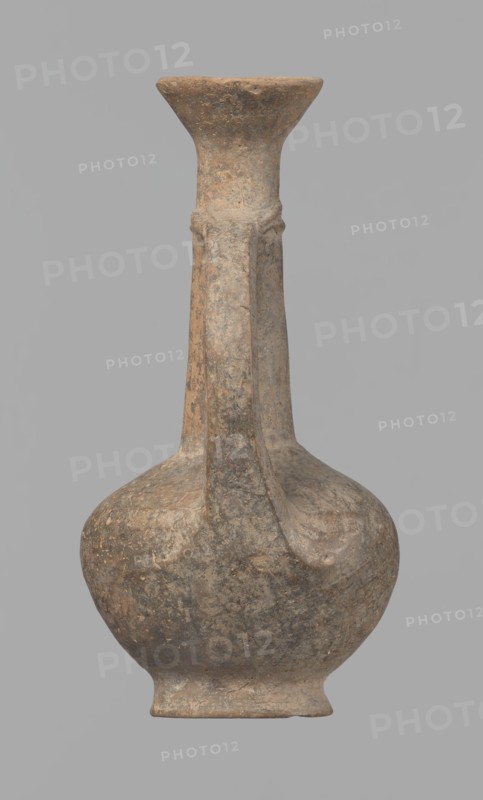Jug, Cypriot pottery, Base Ring Ware I, 1600–1450 BC. Jug with an echinus-shaped foot; a biconical, slightly rounded belly; a high, narrow neck terminating in a funnel-shaped, flared spout. The neck is set at a slight inclination from the plane of the foot. A ribbon-shaped handle is mounted two-thirds up the neck and to the arms of the vessel; at the neck it merges into a double applied ring surrounding the neck. Pinkish clay with a brown surface, in places fading to a brick-red hue. Surface heavily rubbed with slight loss of the rings on the neck. Origin: Cyprus. Vessel; height 12.3 cm, diameter 6.7 cm., An aged clay vessel with a rounded body, a narrow neck that flares into a wide rim, and an attached vertical handle; the surface shows mottled earthy tones including brown, tan, gray, and hints of darker speckling, with a weathered, textured appearance and areas of uneven coloration.