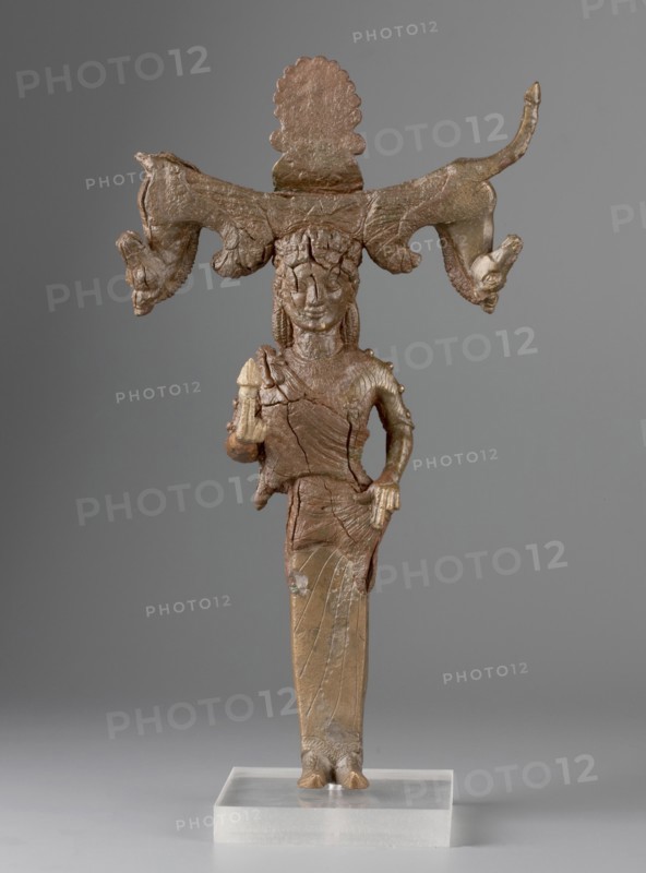 Mirror support with a depiction of the goddess Aphrodite. Support for a standing mirror; hand mirror; figurine of Aphrodite wearing an Ionian chiton and himation. Greek deity depiction, ca. 540–550 BC attributions: Zdrojewska places it on Samos ca. 540 BC; Franck and Furtwängler on the Peloponnese or in Arcadia 550–530 BC; Wallenstein in Corinth, dated to 550 BC. Samos place of origin. Mirror, height 23.2 cm, width 14 cm, depth 3 cm. Maker, inscription/author unknown., A standing bronze figurine with a brownish-green patina mounted on a clear rectangular base, depicting a humanlike figure wearing a long skirt and adorned with a large horizontal headdress that extends outward on both sides and ends in animallike heads; the figure has stylized facial features and textured hair, one hand resting near the hip while the other holds a small object, the surface shows cracks and uneven texture, and the background is a smooth gray.