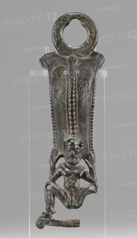 Situla handle with a depiction of Silenus; unknown creator; Silenus mythology; bronze vessel; 1st half of the 5th century BC; Etruria; vessel; height 13.7 cm; width 4.2 cm, The image shows a vertical metal object with a patinated surface in shades of green, brown, and dark gray, mounted against a neutral gray background. The top of the object is rounded with a circular opening and a textured rim. The central section tapers downward and features a raised vertical ridge decorated with a row of bead-like protrusions and bordered by parallel dotted edges. Below this, a small sculpted humanoid figure is integrated into the lower portion, depicted with a bearded face, distinct facial features, a bare torso, and bent legs forming a squatting posture with feet and lower limbs extending outward. The figure appears attached to the broader upper section by its raised arms and sides, and the lower edge of the object includes a short horizontal extension with a curved end. The object rests on a tra...