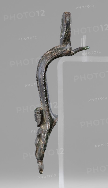 Situla handle with a depiction of Silenus; maker, Silenus mythology; bronze vessel; 1st half of the 5th century BC; Etruria; vessel; height 13.7 cm; width 4.2 cm., A vertical metal object with a curved upper loop and an elongated S-shaped body is shown against a plain gray background; the object has a dark metallic surface with areas of greenish patina and brownish discoloration, a beaded raised ridge running along the outer curve, and a small sculpted human-like figure attached to the lower portion with a distinct head, facial features, and a torso; the piece rests on a translucent clear support with a few clear pegs visible.