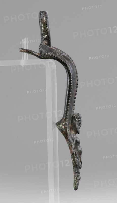 Situla handle with a depiction of Silenus, Silenus mythology, bronze, 1st half of the 5th century BC, Etruria, vessel handle, height 13.7 cm, width 4.2 cm, A curved metal object with a dark brown to greenish patina is shown against a plain gray background, mounted on a clear acrylic stand; the metal surface has areas of green corrosion and darker brown spots, and a row of raised bead-like textures runs along the inner curve; near the lower end a small sculpted human figure in profile is integrated into the form, its surface showing similar brown and green tones and worn detailing on the face and torso, while the upper portion of the object bends into a loop with mottled dark and greenish coloration.