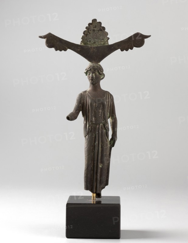 Mirror support with a depiction of Aphrodite. Aphrodite myth., Greek deity. Disc support for a standing mirror; figurine of Aphrodite wearing a Doric chiton. Ca. 450 BC, Sicyon Sykyonia. Hand mirror, mirror figurine. Height 15.5 cm., A small bronze figurine of a standing person mounted on a dark rectangular base, shown against a light gray background; the figure wears a long draped garment with visible folds, has short wavy hair, and one arm extended forward while the other rests at the side; perched atop the figure's head is a horizontal decorative element shaped like a wide V with curled ends and a scalloped piece rising in the center; the object shows areas of greenish patina and overall dark brown metal tones, with the base appearing black and the background light gray.