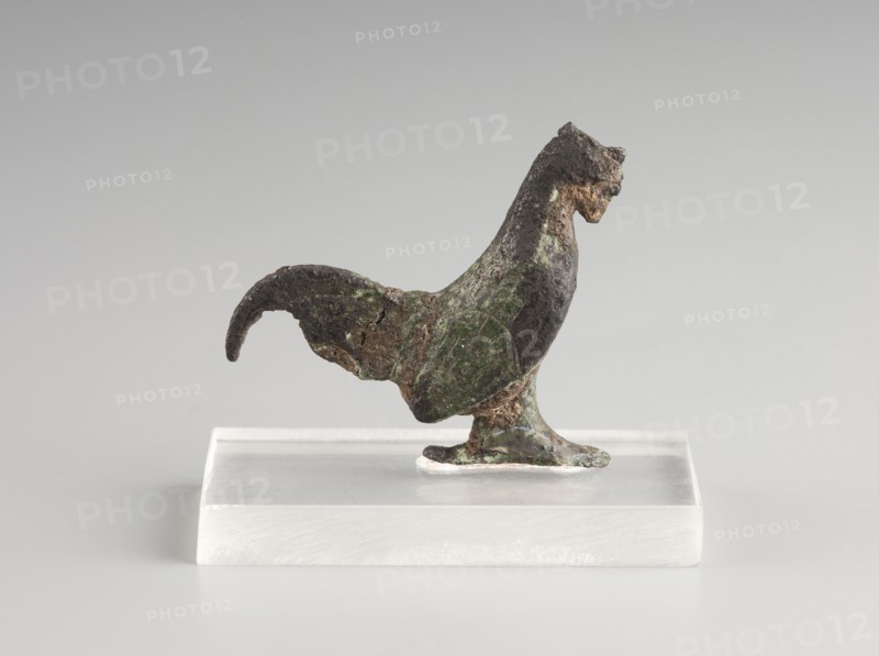 Mirror decoration, 1st half of the 5th century BC, Corinth, mirror, height 3 cm, width 3.6 cm, A small sculptural object of a bird shape stands on a transparent rectangular base; the bird has a curved tail, an upright neck, and a textured surface showing areas of dark brown, green, and hints of lighter brown, with rough, uneven edges and patches of corrosion; the base is translucent and pale, and the background is a smooth, light gray.