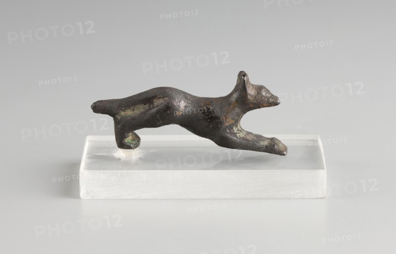 Mirror decoration, unknown author, 1st half of the 5th century BC, Corinth, mirror, height 1.8 cm, width 3.6 cm, A small dark metallic sculpture of an animal in a leaping pose rests on a translucent rectangular base; the sculpture shows a rough, patinated surface with areas of greenish and brownish discoloration and lighter spots, and the base appears white to clear with a smooth finish against a light gray background.