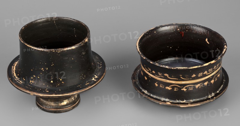 Black-finned pyxis decorated with vine, Greek ceramics, plant motifs, 275–250 BC, Athens, Attica, pyxis vessel, weight 0.16 kg, height 8.3 cm, diameter 8.5 cm., Two shallow cylindrical vessels sit on a smooth gray surface, each with a flared rim and pedestal base; both are primarily black with worn areas revealing a tan or beige underlayer, and one of them displays decorative bands of tan floral and vine motifs around its exterior. The left vessel has a wider flared top with scattered tan speckles and edge wear, a slightly tapered body, and a short rounded pedestal that shows patched tan wear, while the right vessel has a more upright cylindrical profile with two horizontal tan bands and a pattern of small floral shapes and curved lines, along with chipped edges and visible interior wear. The background is a uniform mid-gray.