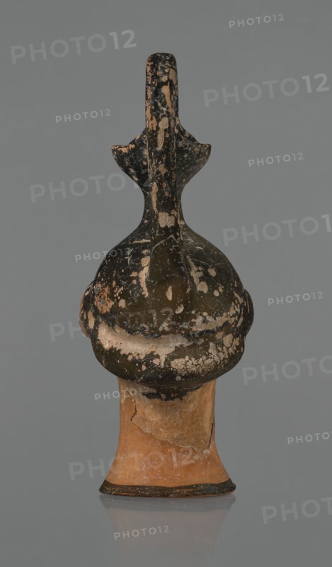 Oinochoe in the shape of a woman's head, repossession Bytom, Greece, oinochoe, head iconography, Greek culture, ancient vessels, yellowish clay, orange surface, in places shiny black engobe. Some elements painted white. Oinochoe in the shape of a woman's head. The upper part is covered with black engobe, as is the headdress, on which a white olive wreath stands out. The neck is outlined at the base with a black band. The elongated female face, very carefully modeled, corresponds to the classic Attic head type from the second half of the 5th century. The oval eyes are surrounded by a black outline. The hair is divided into two parts by a parting and falls in waves to ear level. The headdress flows into a narrow neck ending in a wide, cup-shaped spout. A cylindrical vice is attached vertically between the rim and the back of the head. Preservation condition: missing engobe in some places, damaged surface. Ca. 450 BC, Attica, oinochoe, vessel, An upright ceramic object with a rounded, bul...