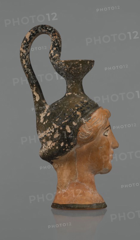 Oinochoe in the shape of a woman's head. Yellowish clay with orange surface and areas of shiny black engobe; some elements painted white. The upper part and headdress are covered with black engobe, with a white olive wreath on the headdress. The neck is outlined at the base with a black band. The elongated female face, very carefully modeled, corresponds to the classic Attic head type from the second half of the 5th century. Oval eyes are surrounded by a black outline. Hair is parted and falls in waves to ear level. The headdress flows into a narrow neck ending in a wide, cup-shaped spout. A cylindrical handle vice is attached vertically between the rim and the back of the head. Preservation: missing engobe in places, damaged surface. Ca. 450 BC, Attica. Oinochoe vessel., The image shows a sculpted head-shaped vessel photographed in profile against a plain gray background, with the face turned to the right. The object combines a terracotta-colored human head with a dark vessel forming ...