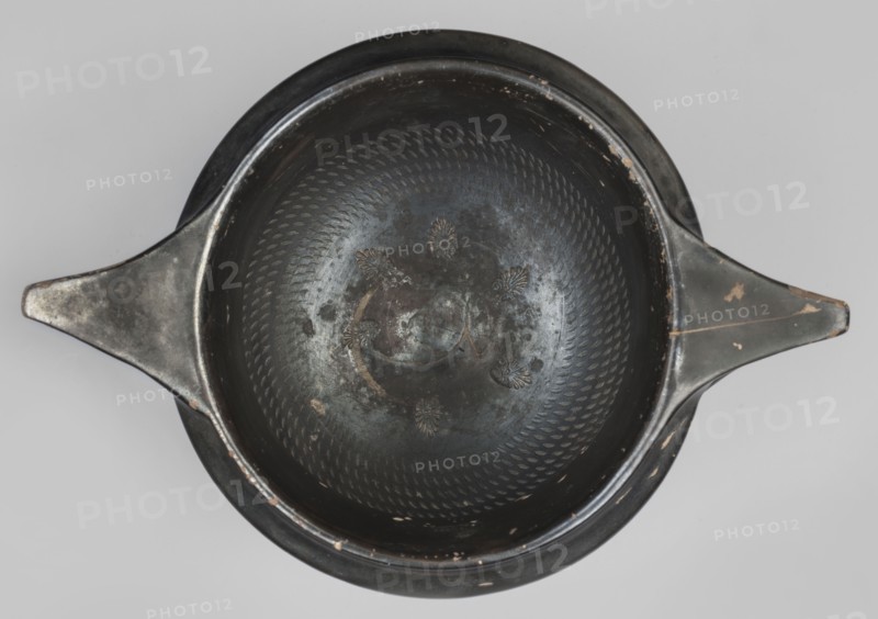 Kantharos, unknown Attic workshop; 320–310 BC, Attica. Kantharos vessel, weight 0.692 kg. The vessel has a high, echinus-shaped foot modeled with a single recess at 3/4 of its height. Rounded, fluted body under the vises the recesses take an oblique direction, creating a three-leaf pattern — on one side the side vises are ajar due to lack of space, narrow shoulders, and a high, smooth neck. The vises are strongly rounded. Modeled spout with a strip below the rim, and characteristic horizontal, elongated, triangular, slightly raised handles above the vises. The bottom features an ornament of six 13-leaf palmettes, not entirely symmetrically arranged; above is a roulette-like decoration 6 rows. The entire vessel, including the inner surface of the foot, is covered with varnish except for the edge of the foot on which the vessel stands and a single depression on the foot. Technology: varnish is shiny, rather metallic, distributed evenly over the surface. Preservation: a few chips, abrasio...
