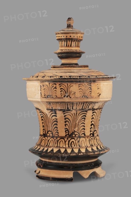 Pyxis with a lid, unknown Boeotian workshop. Pyksis with a lid with a high handle. Cylindrical body with slightly concave walls. Decorated with a plant motif. Set on three legs. Sharp edge above the leg. Bottom decorated with a black circle, with a varnish stain in the central part. Technology: visible traces of turning, good quality glossy varnish, cleaned clay. Notes: reconstructed artefact, large part of the rim and body of the vessel and lid, plaster additions. Maker attribution/author unknown. 2nd half of the 4th century BC, Boeotia. Type: pyxis vessel. Weight 0.950 kg., A ceramic lidded vessel with a rounded base and a tall urn-like top, shown against a plain gray background, featuring a beige or light tan body decorated with black painted ornamental patterns including repeated curved and feather-like motifs, bands, and triangular shapes; the lid has concentric horizontal ridges and dotted and striped black accents, and the base shows chipped areas and wear with some missing frag...