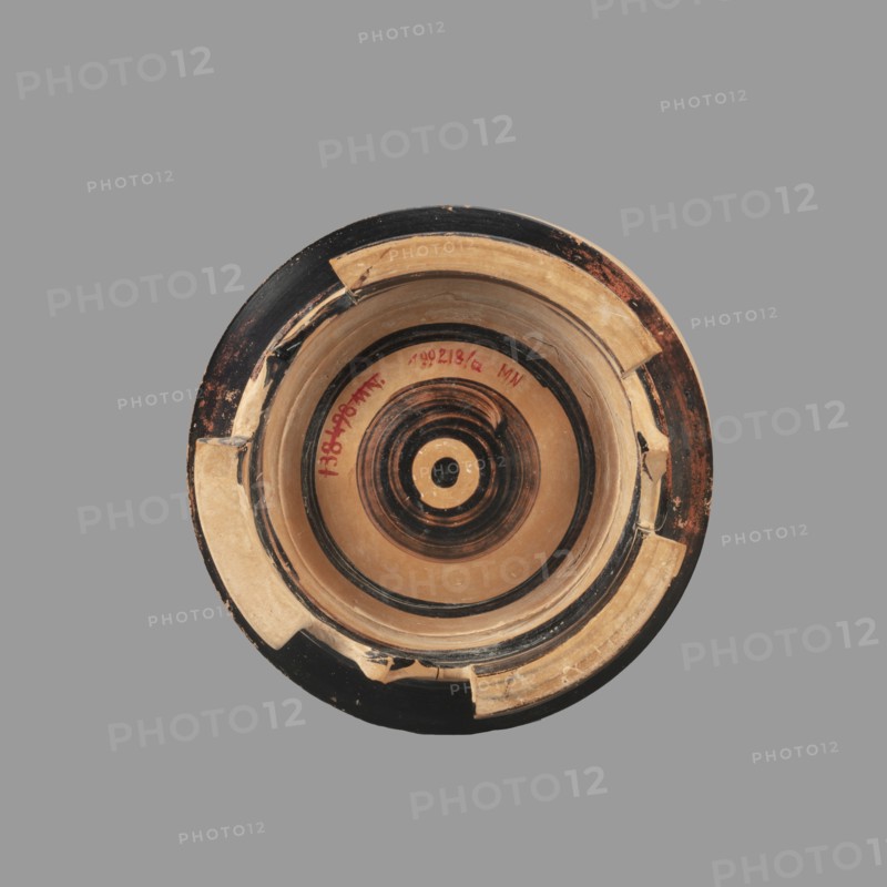Pyxis with a lid, unknown Boeotian workshop. Pyxis with a lid with a high handle. Cylindrical body with slightly concave walls. Decorated with a plant motif. Set on three legs; there is a sharp edge above the leg. The bottom is decorated with a black circle with a varnish stain in the central part. Technology: visible traces of turning; good quality glossy varnish; cleaned clay. Notes: reconstructed artifact—large part of the rim and body of the vessel and lid have plaster additions. 2nd half of the 4th century BC, Boeotia. Pyxis, vessel; weight 0.950 kg., A circular ceramic object viewed from above with concentric rings and a central circular motif, showing areas of tan, black, and dark brown; the rim has chipped and repaired sections with lighter tan patches and visible wear, and the background is a uniform gray.