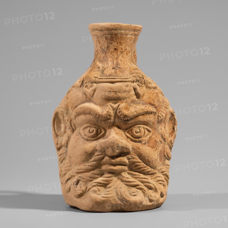 Vessel with a belly in the shape of a satyr's head, unknown Pergamon workshop. A flat-bottomed vessel with a plastic belly in the shape of a Silenus's head, a funnel-shaped spout, and a vertical ribbon handle curved into a circle. The Silenus's head has horse ears and an ivy wreath with flowers above the forehead. The face has a low, wavy cola, small eyes with thin, slender eyelids and marked irises and pupils, a short, broad nose, chubby cheeks, and a narrow, closed mouth. A significant part of the face is covered by a beard and mustache, parted in the middle and arranged in twisted locks with curled ends. Orange clay, 2nd century, Pergamon; vessel. Height 13.6 cm, width 9.6 cm, depth 10.5 cm, weight 0.246 kg., A terracotta vessel shaped like a stylized human head with a pronounced brow, deep-set round eyes, a broad nose, and a full curled beard; the surface shows textured sculpted details and worn areas. The object sits on a flat surface and casts a soft shadow. The background is a s...