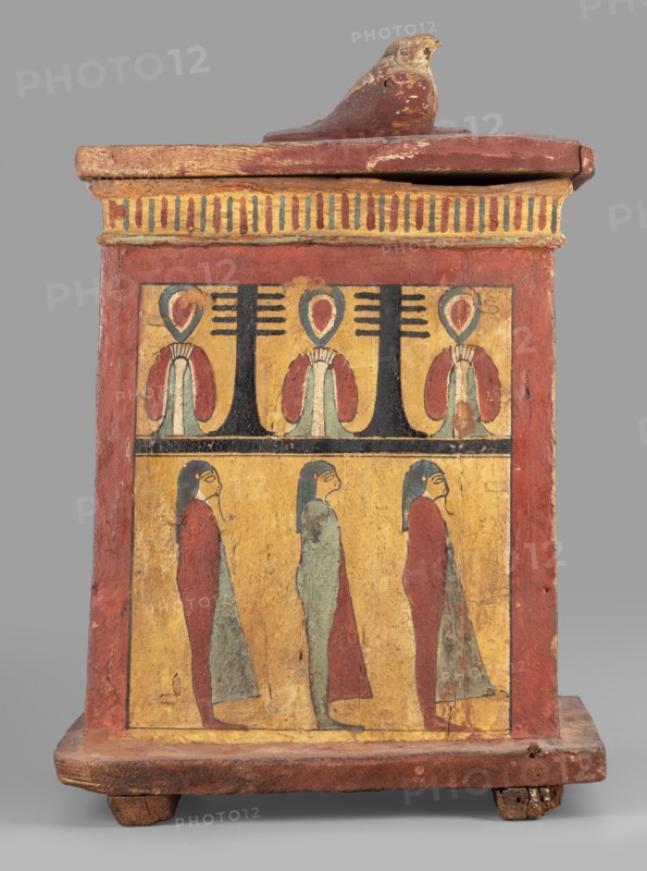 Canopic box with the falcon Sokaris on the roof; falcons; djed pillars iconographic; tit iconographic; naos; grave goods; 664–525 BC, Late Period, 26th Dynasty, Egypt; canopic box; height 42 cm, width 22.5 cm, depth 29.5 cm, weight 2.414 kg;, A rectangular wooden box with a lid topped by a carved bird figure sits on a short base with small feet; the box is painted in muted red, yellow, black, blue-green, and pale green tones. The front face is divided into two horizontal registers bordered by a red frame: the upper register shows a repeated decorative motif with stylized plant-like shapes in yellow and black and teardrop elements in red and blue-green, while the lower register depicts a group of standing human-like figures rendered in profile wearing long garments in red and blue-green against a yellow background. The lid and upper edge display a striped decorative band in alternating yellow and darker hues, and the wooden surfaces show areas of wear and chipped paint.
