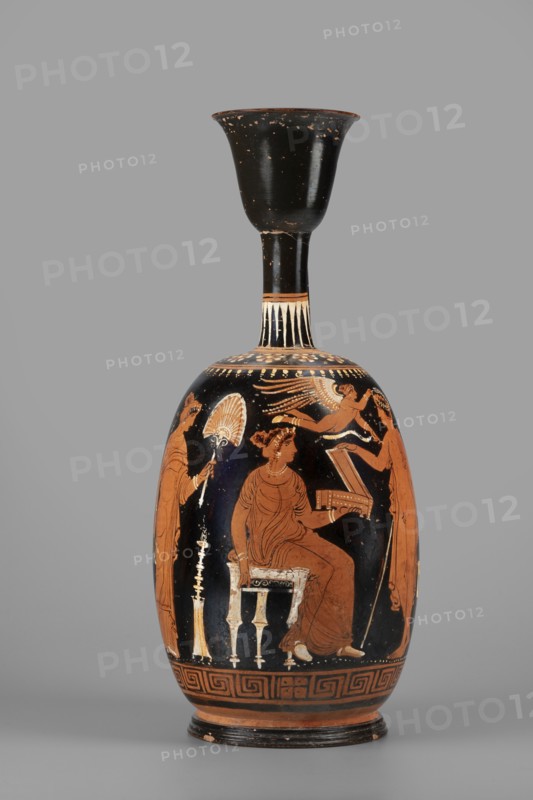 Ariballus lekythos with a depiction of a seated woman, unknown Apulian workshop, Classified Gallo-Classical Scenario, Eros mythology, Greek deities, Southern Italian ceramics, Seated woman with a casket. On the sides a youth crowned by Eros and the figure of a woman. 350–325 BC, Apulia, Ariballus lekythos, vessel, height 28.4 cm, width 12.3 cm, weight 0.520 kg, A tall, narrow ceramic vessel with a black neck and flared rim and a rounded body decorated in orange and black tones, showing a seated woman in an orange dress on a small stool, a standing figure holding a fan to her left, another standing figure to her right holding an object, and a small winged figure above; decorative bands with geometric and floral motifs encircle the shoulder and base, and a few lighter cream-colored highlights accent details on the figures and furniture against a gray background.
