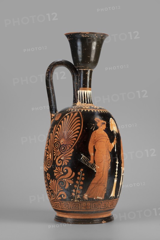 Ariballus lekythos with a depiction of a seated woman; unknown Apulian workshop; Classified Gallo-Classical Scenario; Eros mythology, Greek deities; Southern Italian ceramics; Seated woman with a casket. On the sides a youth crowned by Eros and the figure of a woman. 350–325 BC; Apulia place of finding; Ariballus lekythos vessel; height 28.4 cm, width 12.3 cm, weight 0.520 kg., A tall ceramic vessel with a narrow neck, a flared rim, and a single curved handle; the surface shows black and terracotta tones with areas of wear where the underlying clay shows through. The body features painted decorative motifs in reddish-orange against a black background, including a stylized foliate pattern on one side and a standing robed figure on the other holding a fan-like object, with additional small decorative elements around them. The neck and shoulder have bands of vertical palmette-like strokes and touches of white paint, and the base has a repeating geometric border in reddish-orange and black...