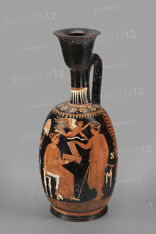 Ariballus lekythos with a depiction of a seated woman, unknown Apulian workshop, Classified Gallo-Classical Scenario. Eros mythology, Greek deities. Southern Italian ceramics. Seated woman with a casket; on the sides a youth crowned by Eros and the figure of a woman. 350–325 BC. Apulia place of finding. Ariballus lekythos vessel; height 28.4 cm, width 12.3 cm, weight 0.520 kg., A tall ceramic vessel with a long neck and flared rim sits against a plain gray background; the vessel is primarily black with areas of reddish-orange and pale cream decoration, showing a painted scene on its rounded body with figures in reddish-orange silhouettes detailed with cream highlights. The neck and handle are black with small worn spots revealing lighter undercolor, and a band of pale cream vertical strokes encircles the neck base. The main painted scene includes a seated robed figure on a small stool holding an open box, a standing robed figure holding a staff and reaching toward the seated figure, an...