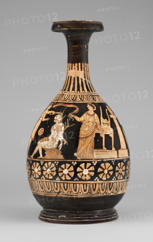 Red-figure bottle with a scene of the punishment of Marsyas, Between the ornamental bands covering the lower half of the neck and belly, a scene depicts the punishment of Marsyas. Marsyas sits on the field of a cloak, the other end covering his right thigh. The lower part of his torso faces left, while the upper part, with his hands tied to a tree in front, faces right toward Apollo standing next to him. Apollo stands beside a table on a platform, holding a lyre placed on it, and in his right hand a taenia, which flutters above Silenus's head. A Scythian stands before Marsyas, using a knife held in his right hand, and begins to flay him. The scene is complemented by three satyrs and a column with an oval object possibly an omphalos. The scene reflects the musical contest agon between Apollo lyre and Marsyas aulos. Red-figure Greek pottery, 330–310 BC. Bottle vessel. Height 26 cm, width 13.5 cm, weight 0.67 kg. Vessel glued from several fragments; minor additions on the neck; flaking of...
