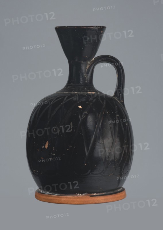 Ariballus lekythos, 4th century BC, Nola findspot, Campania origin, lekythos vessel, height 12.0 cm, width 7.0 cm, A dark vase with a bulbous body, a narrow neck flaring into a wide mouth, and a single curved handle at the shoulder, sitting on a flat round base; the vase surface is predominantly black with visible scuffs and small areas where the dark coating is worn away to reveal a lighter material, and the base ring is a terracotta color; the background is a flat muted gray.