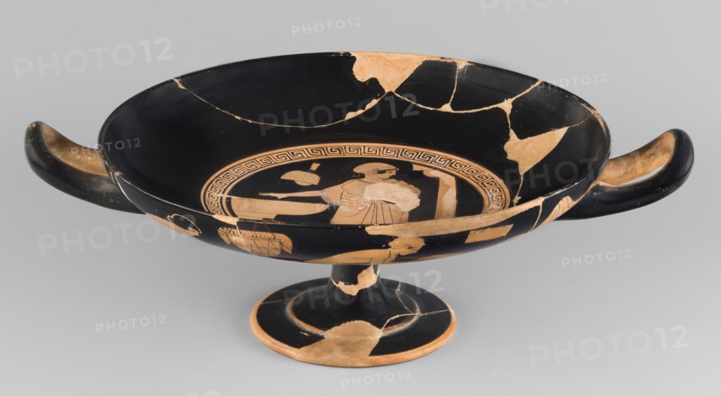 Red-figure kylix with a bathing scene, the Boots Painter ca. 460 BC. Bath iconography; Greek ceramics. Physical description: goblet with slightly parted rim edges, shallow bowl, low stem, and flat foot. Vices have a round cross-section, slightly raised tops reaching the line of the rim. Conical bottom. Sleeve-shaped foot. Scene: Women wash at lutrions bowls on high bases, pass around folded garments, and one of them ties characteristic high boots. Technique: Thick, very good quality varnish, evenly distributed; smooth surface; traces of turning invisible, brush marks visible. Mixture: mica. Condition notes: Artifact glued from many fragments with visible conservation restorations; surface contaminated; visible secondary losses and chips; standing surface heavily worn. Attributed to Kamila Nocon; provenance/attribution notes: Attica Athens, Tarquinia Beazley 1928. Kylix; vessel. Dimensions: height 9.2 cm, width 29.8 cm, depth 22.8 cm, weight 0.53 kg., A shallow, wide ceramic bowl with a...