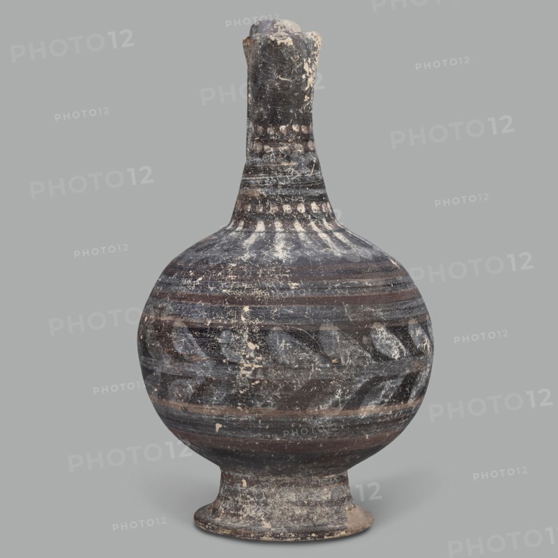 Oinochoe, unknown Cypriot workshop; Arsinoe Cyprus. Oinochoe on a cup-shaped foot with a spherical body transitioning into sloping arms, separated by a recess from the narrow, tapering neck terminating in a triangular rim. The band-shaped handle protrudes slightly above the rim and is connected to the arm. The vessel is covered with gray-blue, matte paint. Decoration in white and brown paint surrounds the vessel concentrically. On the foot, stripes of white paint decorate the body; a laurel sprig adorns the narrowest part of the body, with white and brown stripes above it. On the shoulders, a ray ornament extends up to the neck. On the neck at the base and under the rim, white dots, and between the rows of dots, white stripes. 332–30 BC; Hellenistic period, Cyprus. Oinochoe vessel, height 14 cm, width 9 cm., A rounded ceramic vessel with a narrow neck and flared foot, shown against a plain gray background. The surface of the vessel displays worn horizontal bands and decorative patterns...