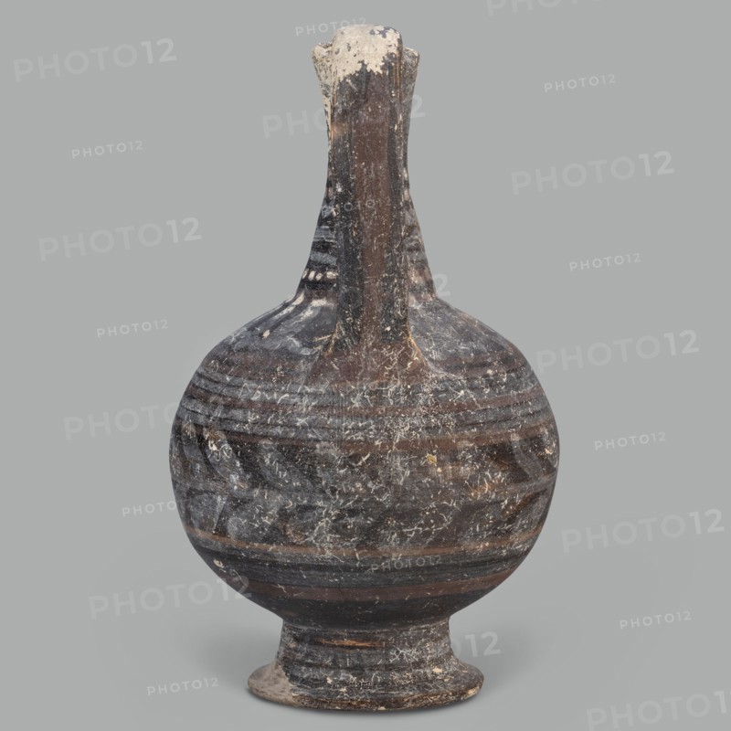 Oinochoe, unknown Cypriot workshop; Arsinoe Cyprus. Oinochoe on a cup-shaped foot with a spherical body transitioning into sloping arms, separated by a recess from the narrow, tapering neck terminating in a triangular rim. The band-shaped handle protrudes slightly above the rim and is connected to the arm. The vessel is covered with gray-blue, matte paint. Decoration in white and brown paint surrounds the vessel concentrically. On the foot, stripes of white paint decorate the body; a laurel sprig adorns the narrowest part of the body, with white and brown stripes above it. On the shoulders, a ray ornament extends up to the neck. On the neck at the base and under the rim, white dots, and between the rows of dots, white stripes. 332–30 BC; Hellenistic period, Cyprus; oinochoe vessel; height 14 cm, width 9 cm., An aged rounded vessel with a narrow tall neck and a flared foot is shown against a plain gray background; the surface displays a weathered, mottled pattern in dark brown and black...