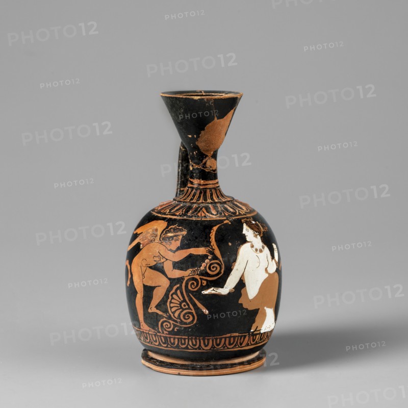 Aryballus lekythos. An aryballus lekythos on a ring-shaped, profiled foot, with a bulging, egg-shaped belly, convex handles flowing into a neck terminating in a cup-shaped rim. The vise is narrow and band-like. At the base of the neck is a bar ornament, and on the handles is an egg-shaped motif with plastic lines in ovals. Below the vise are two upward-facing palmettes, arranged one above the other, decorated with leaves and volutes. The front of the vessel depicts a seated, nude woman wearing a garment covering her legs and hips; she faces left with her head turned right. Behind her is a leaning, winged Eros, standing on his right leg with his left leg raised, holding a necklace in his outstretched hands. The composition is framed from below by an egg-shaped motif. Preservation condition: The neck, vise, and rim are glued; a few chips are visible on the surface. Ca. 420 BC earlier dating: 4th century BC. Apulia. Lekythos aryballus lekythos. Vessel dimensions: height 9.4 cm; width 5.0 ...