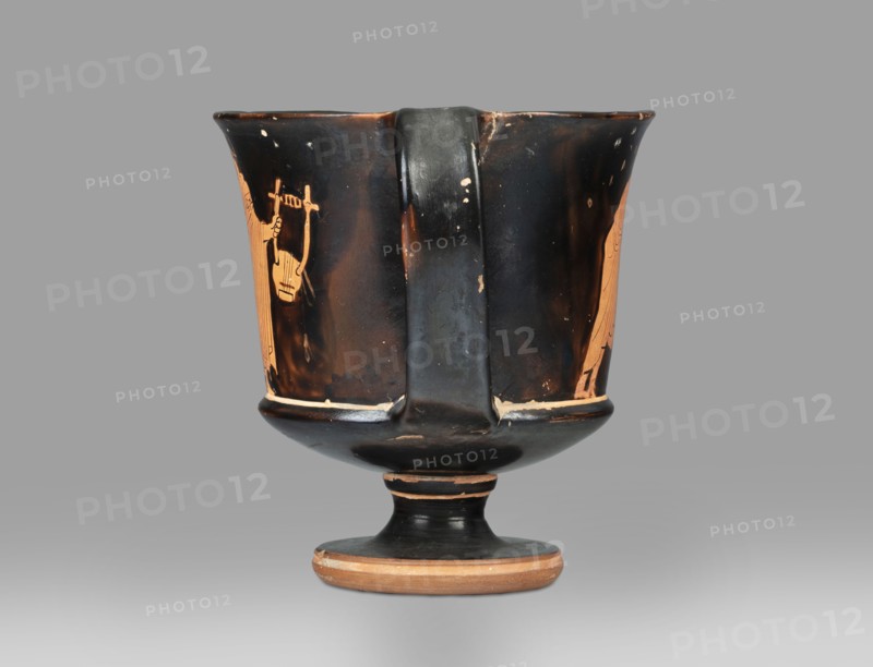 Red-figure kantharos with a depiction of an ephebe with an aryballos and a Greek inscription “Beautiful boy”;, Kantharos with a slightly flared spout, a deep bowl, a flat bottom, a high profiled stem, a ring-shaped foot. Thin-walled. A: bearded man with a lyre in his head in left profile, holding a lyre in his left hand. B: ephebe with aryballos. The inner surface of the bowl is covered with varnish. Technology: Smooth surface. The varnish is thick, shiny, unevenly fired. Notes: the vessel is intact; small secondary losses and chips are visible on the surface. Varnish color: 10 YR 2/1 Black. Date: ca. 450 BC mid-5th century BC. Place of origin: Attica. Type: kantharos, vessel. Dimensions: height 11 cm, width 16.2 cm, depth 10.2 cm., A single ceramic vessel with a broad rim, a central vertical handle, and a footed base sits against a plain background; the vessel shows a glossy black surface with areas of orange showing through where the black glaze is worn or intentionally left, includi...
