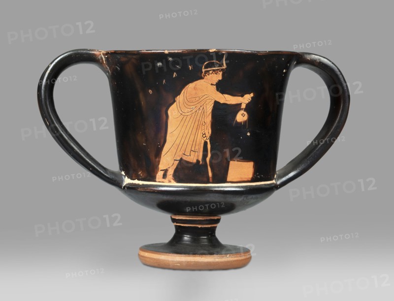 Red-figure kantharos with a depiction of an ephebe with an aryballos and a Greek inscription “Beautiful boy”., Kantharos with a slightly flared rim, a deep bowl, a flat bottom, a high profiled stem, ring-shaped foot; thin-walled. A: bearded man with a lyre in his head in left profile, holding a lyre in his left hand. B: ephebe with aryballos. Inner surface of the bowl coated with varnish. Technique: smooth surface; varnish thick, shiny, unevenly fired. Condition: vessel intact; small secondary losses and chips on surface. Varnish color: 10 YR 2/1 black. Circa 450 BC mid-5th century BC. Attica provenance. Type: kantharos vessel. Dimensions: height 11 cm; width 16.2 cm; depth 10.2 cm., A two-handled ceramic vessel with a rounded bowl and a short stem rests against a neutral background; the vessel has a glossy black surface with orange-red figures and details, showing a standing robed figure in profile reaching toward a small round object held in the other hand, and a rectangular block ne...
