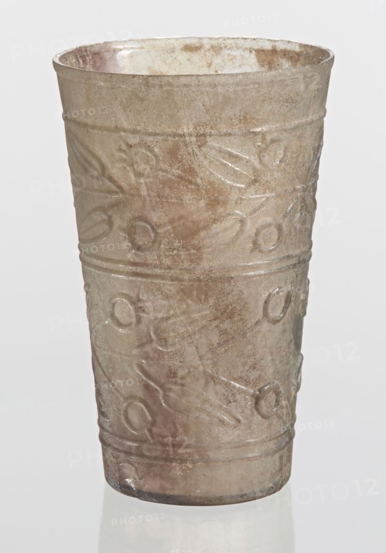 Glass decorated with a leaf and fruit motif, unknown creator; Goluchów Greater Poland Voivodeship; glass vessel with plant motifs; transparent, colorless glass; relief decoration in two registers separated by two horizontal thin convex lines; upper register bounded from the top by a single convex line; lower register bounded from the bottom by two convex horizontal lines; blown in a mold. Bottom diameter: 4.25 cm; dated 1st–2nd century previously dated 2nd century BC; manufactured in Syria; found in Gaul France; vessel; weight 0.066 kg., A tall cylindrical cup with a slightly flared rim, made of translucent glass showing a weathered, iridescent surface with shades of pale brown, gray, and subtle green and pink tones; the exterior features a repeating raised pattern of circular and linear motifs arranged in horizontal bands, with a worn and pitted texture and areas of cloudiness and spotting, and the cup sits on a reflective surface against a light neutral background.