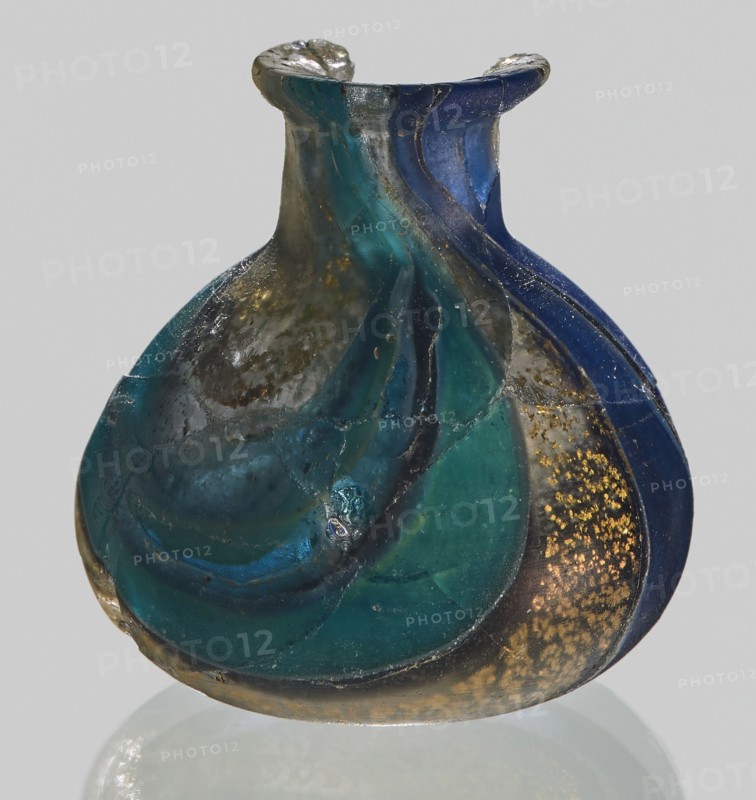 Unguentarium with carnation with a gold ribbon; with a bulging belly. 1st half of the 1st century BC. Type: Isings form 7. Mosaic glass small bottle with a cylindrical neck; sagging. Height: 2.9 cm; Body diameter: 2.6 cm; Weight: 0.008 kg. Unknown workshop; possibly Egyptian workshop, and unknown Italian workshop ., A small rounded glass vessel with a flared neck and a wavy, ribbed surface, showing swirling bands of deep blue, teal, green, and dark turquoise glass, with areas of cloudy translucent gray and patches of golden speckling on one side; the glass surface shows irregularities, seams, and small chips, and the object casts a faint reflection on a neutral light-gray background.