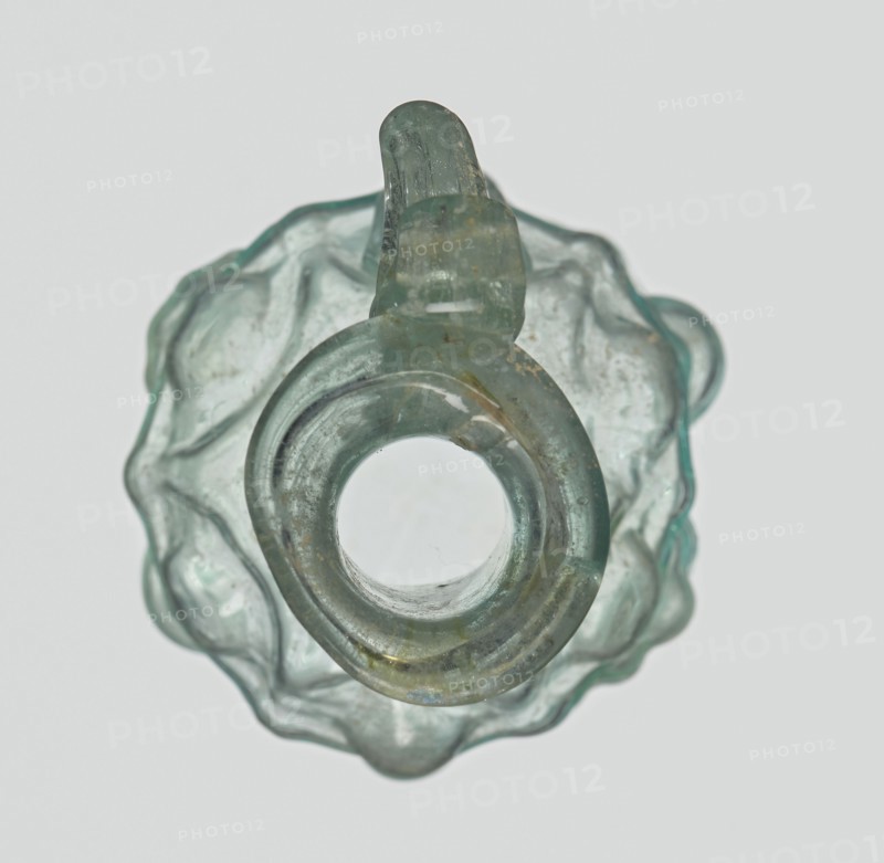 Hexagonal jug, eastern workshop, Syria country of find, 1st century AD. Stern type with vessels, series A. Transparent light green glass; ovoid hexagonal body decorated with a convex relief depicting vessels on the arms of an arcade with convex elements. Cylindrical neck terminating in a flaring spout with an inwardly curved edge; blown in a three-part mold. Outer diameter of the spout: 1.90 cm; height: 9.30 cm; body diameter: 4 cm; outer diameter of the bottom: 2.50 cm; weight: 0.034 kg., A translucent object with a pale green and aqua tint fills the frame, viewed from above showing a circular central opening surrounded by a rounded ring and an irregular scalloped outer edge; the glass-like material has cloudy areas, surface imperfections, and a few small brownish spots, and the overall shape includes a short looped protrusion at the top with similar greenish-blue coloration.
