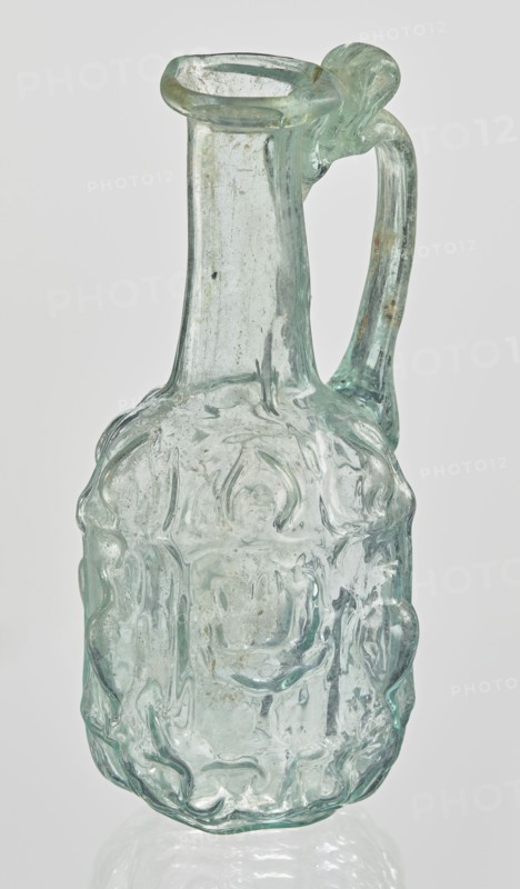 Hexagonal jug, Goluchów Greater Poland Voivodeship. Hexagonal jug; eastern workshop; Syria country of find; 1st century AD. Type: Stern type with vessels, series A. Transparent, light green glass. Ovoid, hexagonal body decorated with a convex relief with a representation of vessels on the arms of an arcade with convex elements; cylindrical neck terminating in a flaring spout with an inwardly curved edge; blown in a three-part mold. Outer diameter of the spout: 1.90 cm. Height: 9.30 cm. Body diameter: 4 cm. Outer diameter of the bottom: 2.50 cm. 9.3 x 4 cm, weight 0.034 kg., A translucent glass pitcher with a greenish-blue tint and irregular, wavy surface texture, showing a narrow neck that flares into a rounded lip, a curved handle attached near the top and middle, and a bulbous body with embossed, flowing raised patterns and visible tiny bubbles and surface imperfections; the background is a plain light gray.