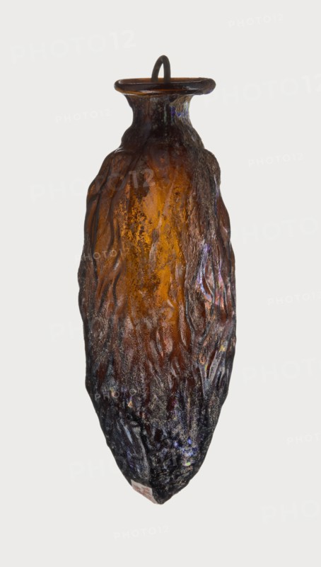 Date-shaped unguentarium; Syro-Palestine workshop; Smyrna place of discovery; 1st–beginning of 2nd century AD. Type: Isings form 78D. Translucent amber glass with inclined rim and inwardly curved edge; blown in a two-part mold. Vessel: rim diameter 2 cm, height 8.15 cm, belly diameter 2.95 cm, weight 0.020 kg., A textured, elongated vessel with a narrow neck and flared rim, predominantly amber and brown in color, with darker brown and almost black areas toward the base; the surface shows irregular, flowing ridges and a rough, pitted texture with subtle iridescent highlights in purple and blue on parts of the sides; a looped handle or ring sits at the rim, and the overall shape tapers to a pointed bottom.