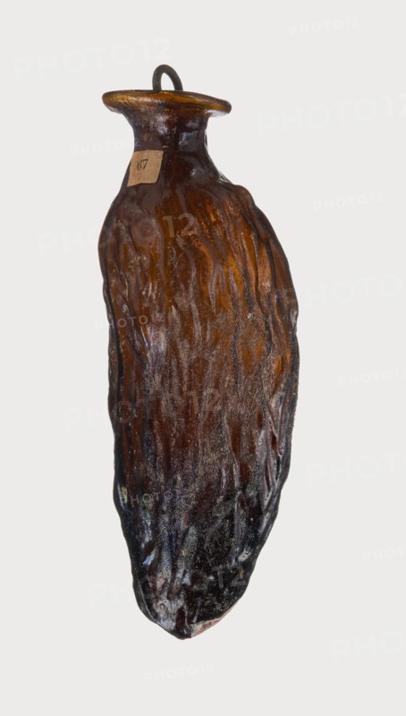Date-shaped unguentarium; Syro-Palestine workshop; Smyrna place of discovery; 1st–beginning of 2nd century AD; Type: Isings form 78D. Translucent amber glass, inclined rim with inwardly curved edge; blown in a two-part mold; 1st century BC–beginning of 2nd century AD. Vessel, rim diameter 2 cm, height 8.15 cm, belly diameter 2.95 cm, weight 0.020 kg., The image shows a single elongated, irregularly shaped object resembling a bottle or vessel with a narrow neck and flared rim, suspended against a pale background. The surface has a rough, textured appearance with vertical ridges and an uneven finish. Visible colors include various shades of brown, amber, dark brown to near-black, and subtle hints of iridescent blues and purples in places. A small loop or handle sits atop the rim in a dark color. The object appears glossy in some areas and matte in others, with lighter amber tones concentrated near the neck and darker tones toward the lower portion.