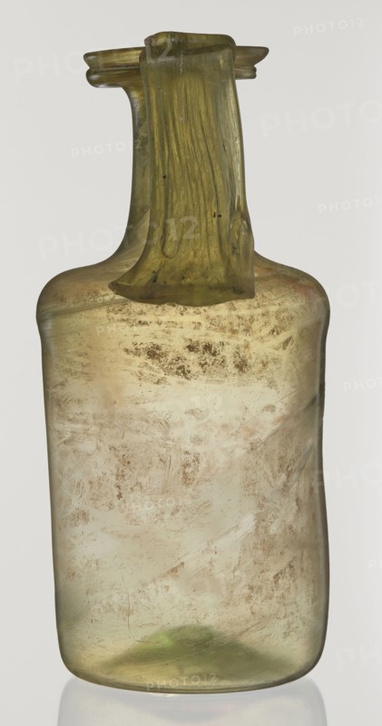 Jug, unknown eastern workshop, cylindrical body, high neck with flared spout and vertical vice, 2nd–3rd century AD, Syria, vessel, height 21.5 cm, width 10.5 cm, A single tall, narrow glass bottle with a rectangular body and rounded shoulders, topped by a short neck and a flattened, wide rim with a draped handle-like strip of glass running from the rim down along the neck; the glass shows a translucent greenish-yellow hue with areas of cloudy, pale beige and brownish surface deposits and mottling, and there are darker green tones near the top and bottom.