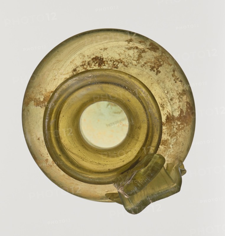 Jug, unknown eastern workshop, Cylindrical body, high neck with flared spout and vertical vice, 2nd–3rd century AD, Syria, vessel, height 21.5 cm, width 10.5 cm, A circular translucent glass object viewed from above with a central round opening, showing a yellow-green color with areas of darker olive and pale green; the surface has mottled brownish spots and patches and a few small dark speckles; a thicker rounded rim surrounds the opening and a folded or crimped glass protrusion extends from one side, matching the yellow-green hue with some internal striations and slightly glossy highlights; the background is a pale off-white.