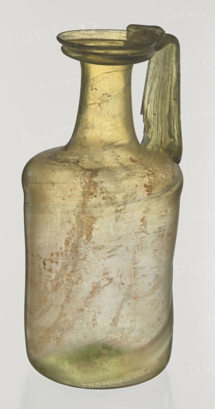 Jug. Cylindrical body, high neck with flared spout and vertical vice. 2nd–3rd century AD. Syria. Vessel. Height 21.5 cm; width 10.5 cm. Maker: unknown eastern workshop., A translucent glass vessel with a narrow neck and flared rim, showing a single handle attached from the rim to the shoulder; the glass is primarily pale yellow-green with areas of cloudy, muted green and brownish weathering; the surface shows irregular patches and streaks of earthy brown and tan discoloration, with a slightly iridescent sheen in places; the handle is a deeper green and appears thicker and slightly irregular in shape; overall the vessel has a worn, aged appearance against a plain light background.