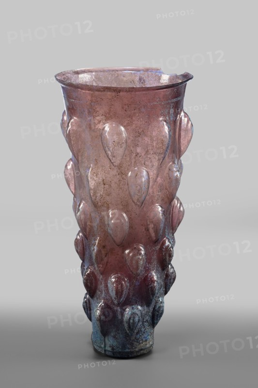 Glass mug; transparent, manganese, iridescent glass. Decorated with 6 rows of convex, stepped elements shaped like almonds lotus buds. Two concentric circles on the bottom; blown in a 4-part form. Isings form 31. Outer diameter of the bottom: 5 cm; weight 0.188 kg. Dated 2nd quarter of the 1st century, Syria., A single tall translucent vessel with a flared rim stands against a plain light gray background, showing a pinkish to purplish overall hue with areas of bluish-green iridescence and weathered discoloration; the surface is decorated with a repeated raised teardrop motif arranged in vertical rows, and the glass shows spots of mottling and patina with darker, more bluish tones concentrated near the base while the upper area remains more pink and slightly worn, and the rim has an irregular small chip.