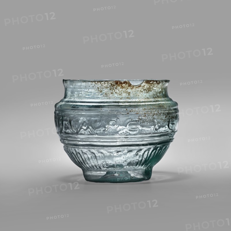 Bowl with Greek inscription. Glass bowl of transparent light blue glass with an uneven rim edge, cold-worked. Relief decoration in the lower register: radially arranged long leaves flutes. In the upper, widest part of the bowl there is a Greek inscription: Rejoice that you have come, bounded at the top and bottom by horizontal ribs. Blown in a three-part form. Type: Harden group G.1.IIa. Date: 1st century manufacture in Sidon; 1st half of the 19th century noted classification. Origin: Sidon; Cyprus discovery. Vessel, weight 0.070 kg; diameter 4.5 cm bottom, diameter 7.4 cm rim, height 7 cm., A translucent glass bowl with a rounded body and a slightly flared rim sits against a neutral gray background; the glass shows a few areas of greenish-blue tint and several patches of brownish discoloration near the rim, with visible air bubbles and irregularities in the surface; the body features a band of embossed decorative motifs and a lower section with vertical ribbing, and the glass rests on...