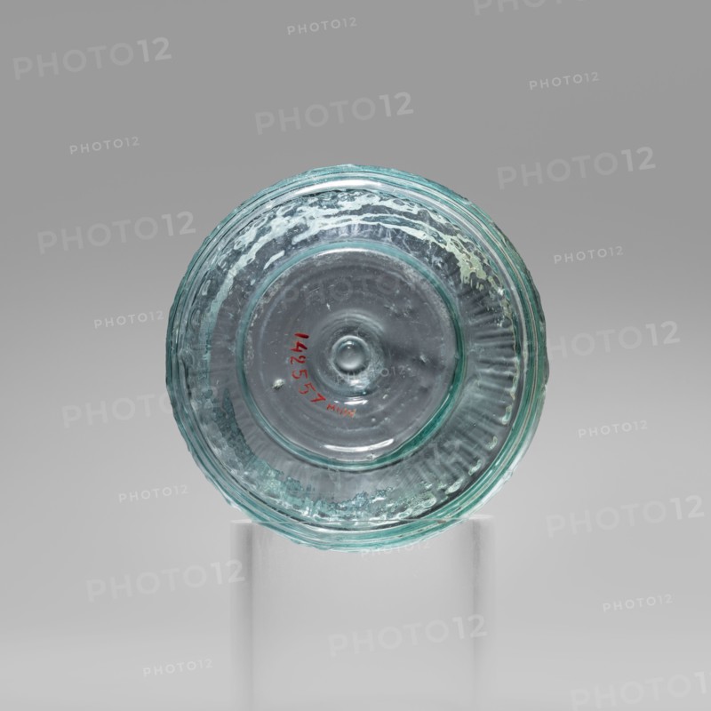 Bowl with Greek inscription. Glass bowl made of transparent, light blue glass with a Greek inscription reading Rejoice that you have come. Uneven rim edge, cold-worked. Relief decoration in the lower register: radially arranged long leaves flutes; inscription in the upper, widest part of the bowl bounded at top and bottom by horizontal ribs. Blown in a three-part form. Harden group G.1.IIa. Dated 1st century Sidon, 1st half of the 19th century note: conflicting dates in original. Origin: Cyprus. Vessel. Weight 0.070 kg; diameter 4.5 cm bottom, diameter 7.4 cm rim; height 7 cm., A round translucent glass object with a bluish-green tint sits centered on a clear cylindrical stand against a neutral gray background; the glass shows concentric circular ridges and a slightly raised central bump, with textured irregularities and small bubbles visible within the material, and a small area of red color near the inner circle.
